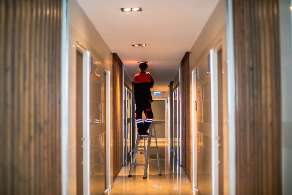 A worker stands on a ladder in a well-lit corridor, fixing a ceiling light. The hallway has reflective floors and wooden paneling on the walls, with doors lining both sides.