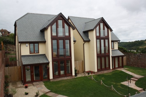 Two adjacent modern houses with large glass windows, cream walls, and dark brown frames. Each house has a lawn and pathways leading to the entrances, all set against a backdrop of cloudy skies and greenery.