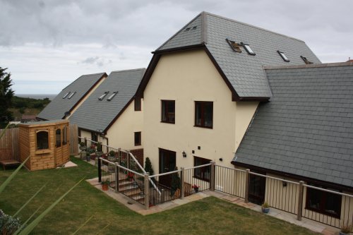 A row of modern, beige houses with dark gray sloped roofs. The houses have multiple windows and skylights. A fenced backyard with a wooden shed is visible, set on a grassy lawn under a cloudy sky.