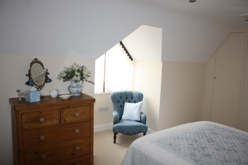 A cozy bedroom with a wooden dresser topped with a mirror, vase with flowers, and white ceramics. A blue armchair with a white pillow sits near a window. The bed has a white quilt, and a slanted ceiling adds a unique touch to the room.