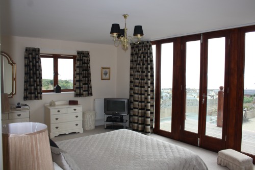 A bedroom with a large bed, patterned curtains, and a wooden dresser. A small TV is placed on a stand in front of a window with a view of a balcony. The room has wooden accents and neutral-colored decor. A ceiling light fixture is in the center.