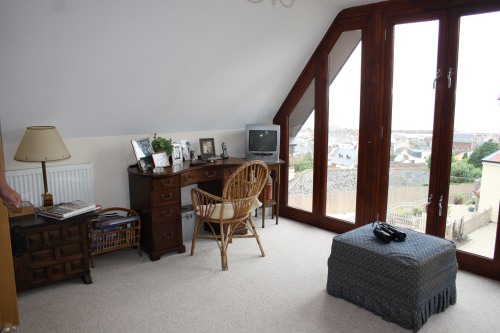 A cozy attic room with a wooden desk and chair near large windows overlooking a town. The desk has a small TV, framed photos, and a lamp. An ottoman and a basket with magazines are nearby on the carpeted floor.