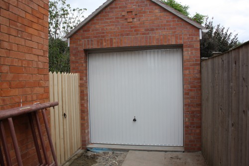 A small brick garage with a closed white door is flanked by a red brick wall on the left and a wooden fence on the right. A wooden ladder leans against the brick wall. Bushes and trees are visible in the background.