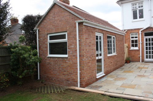 A red brick home extension with a sloped roof, featuring a white-framed window and door. It is attached to a larger white house. The surrounding area has a paved patio and some greenery, including a small tree.