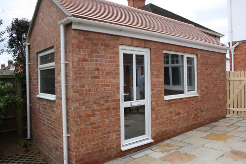 A small brick house extension with a white-trimmed door and two windows. The roof is covered with red tiles. The surrounding area features a paved patio and a wooden fence.