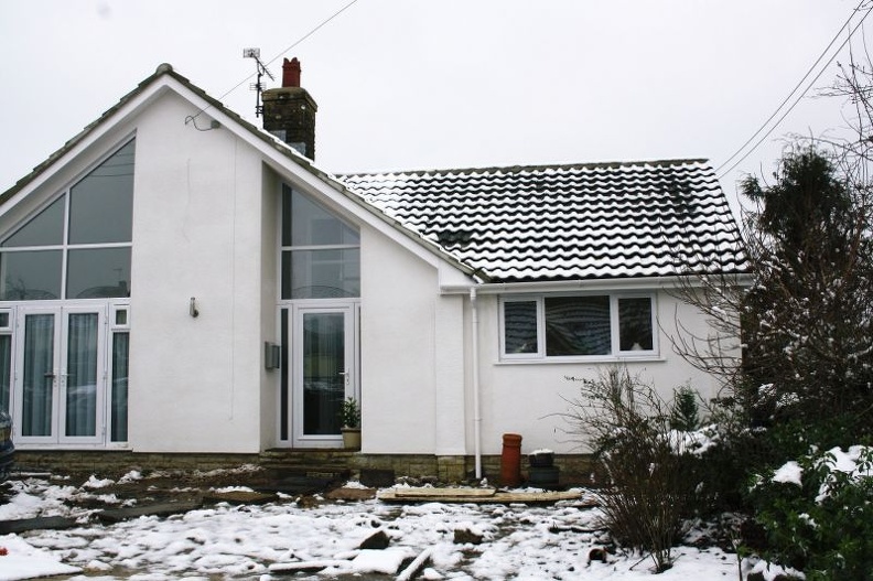 A small, snow-covered house with a gabled roof and large glass windows. The yard is partially covered in snow, with some exposed plants and a clay pot near the entrance. Overcast sky in the background.