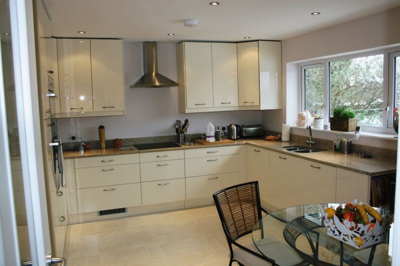 A modern kitchen with cream cabinets, a stainless steel range hood, and a granite countertop. A table with a glass top and a fruit bowl sits in the foreground, next to a large window letting in natural light.