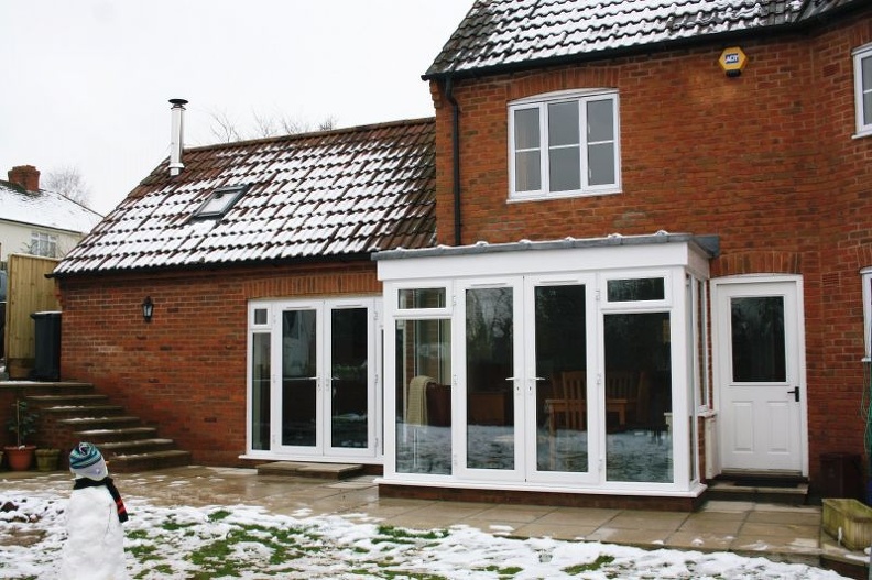 A brick house with a tiled roof features a white-framed conservatory. Snow covers parts of the ground, and a small snowman with a hat stands on the left. A table and chairs are visible inside the conservatory.