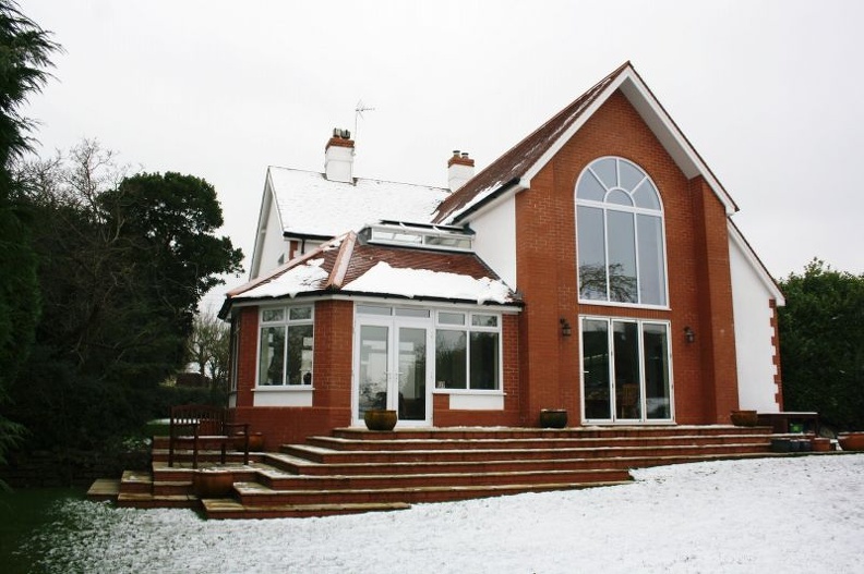 A two-story brick house with a large arched window stands on a snowy lawn. The house has a sloped roof, white-framed windows, and several steps leading to the entrance. Trees are visible on the left side.