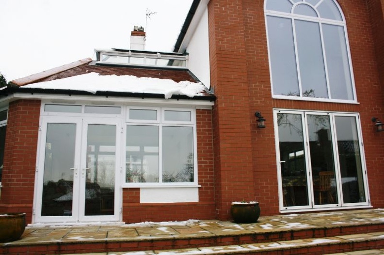 A red-brick house with large windows and a snow-covered roof. The building features a glass double door and adjacent tall windows, set on a stone-paved patio with empty planters.