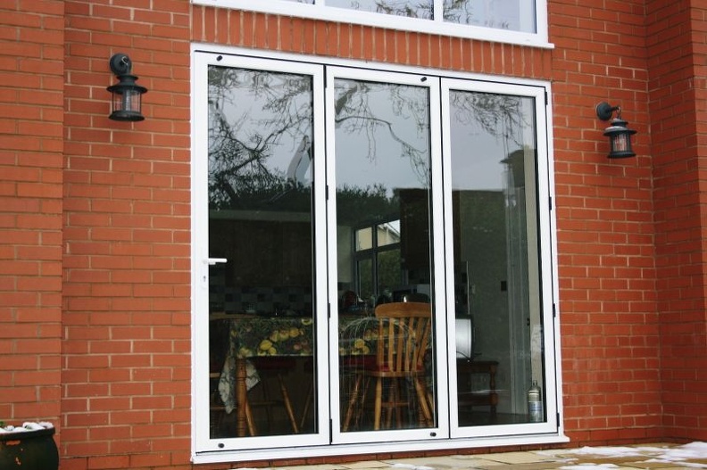 A set of three white-framed glass doors on a brick wall, leading to an indoor area with wooden chairs and a table. Two black wall-mounted lanterns flank the doors. Outside, a small snowy patch is visible on the ground.