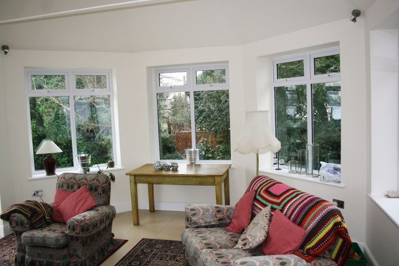 A cozy sunroom with large windows overlooking a garden. It features two patterned armchairs with colorful throws, a wooden table with decor, a standing lamp, and small decorative rugs on the floor. Bright and inviting atmosphere.