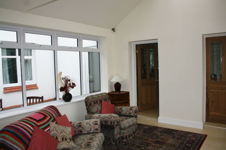 A cozy living room with patterned armchairs and a colorful throw. There are red cushions on the chairs. A small wooden side table with a lamp is next to a large window. The wall and doors are painted white and brown, respectively.