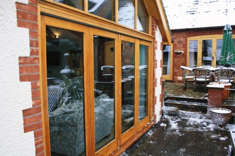 Exterior view of a house with large wooden-framed glass doors leading to a patio. The patio is covered with some snow and has outdoor furniture. A green umbrella is visible, and the surrounding brick and white walls complement the snowy scene.