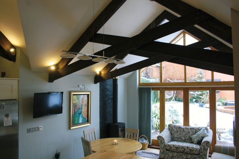 A cozy room with a vaulted ceiling and exposed wooden beams. It features a round dining table, chairs, a patterned couch, a wall-mounted TV, and a large window overlooking a snow-covered patio. The walls are light blue with a framed painting.