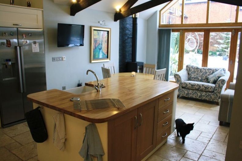 A cozy kitchen with a large wooden island, stainless steel fridge, and mounted TV. A black cat walks on the tile floor. Large windows in the background reveal a snowy patio with outdoor seating.