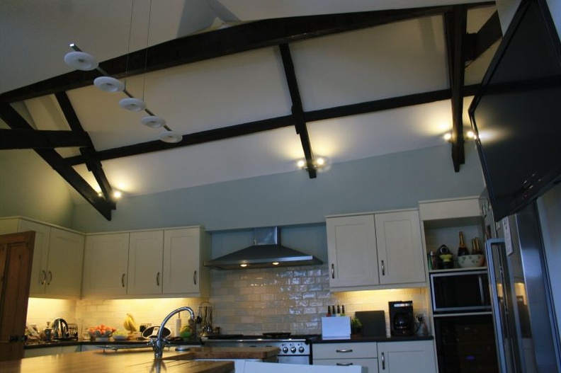 A modern kitchen with cream cabinets, stainless steel appliances, and a sleek range hood. Exposed dark wooden beams add character to the high ceiling. Soft lighting highlights the space, and fruit is displayed on the countertop.