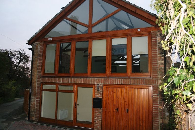 Two-story brick building with large wooden-framed windows. The upper level has several triangular and rectangular panes, while the lower level features a series of glass doors. There is also a wooden garage door on the right. Vegetation is on the side.