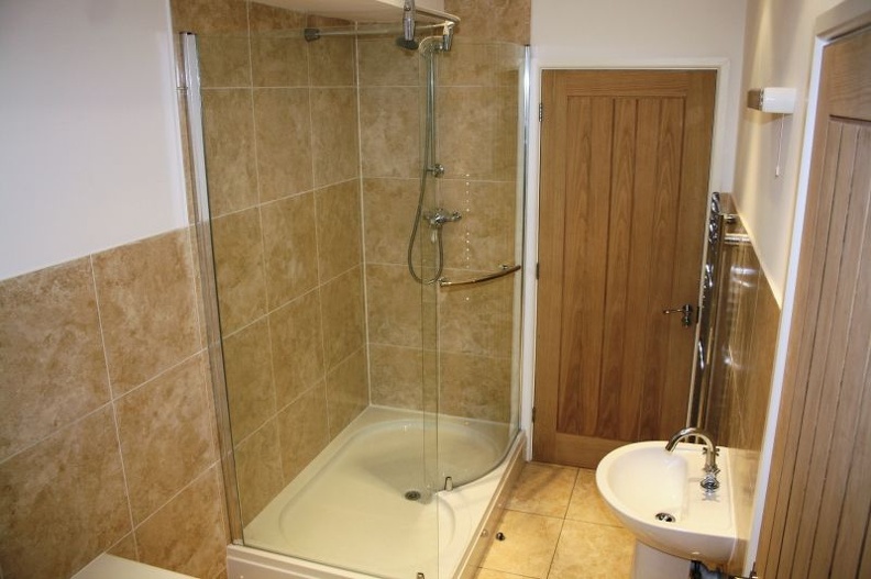 A bathroom with a glass-enclosed corner shower featuring beige tiles. There is a wooden door on the right and a white sink with a silver faucet below a wall-mounted mirror. The floor is tiled in a light beige color.