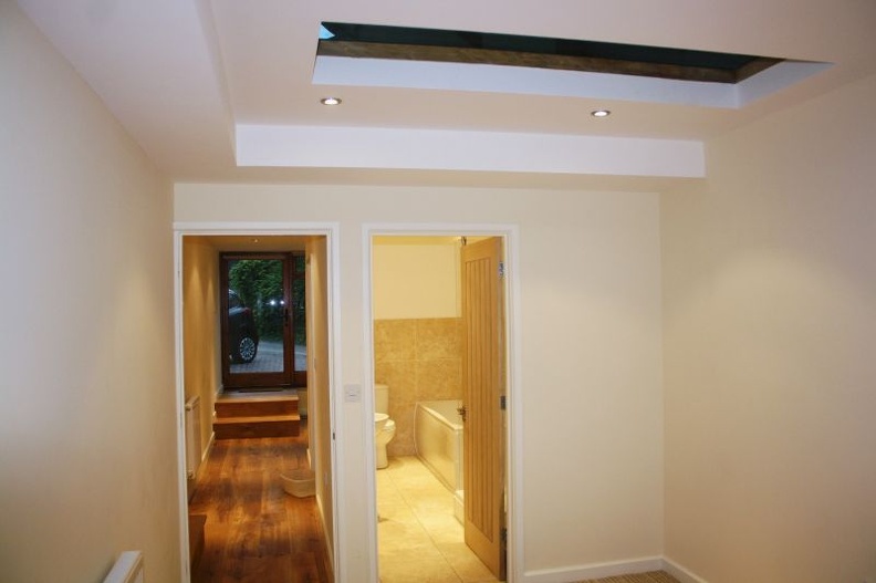 Interior hallway with a skylight, leading to a wooden-floored room on the left and a bathroom with tiled floors and a bathtub on the right. Walls are painted white, and there are recessed ceiling lights.