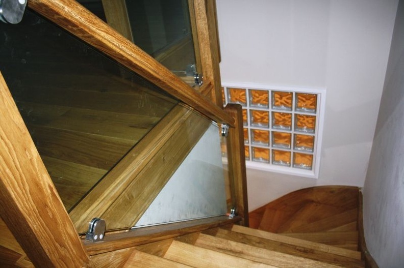 A modern spiral staircase with wooden steps and glass panels, descending alongside a wall with glass blocks that allow light to pass through, creating a warm and welcoming atmosphere.