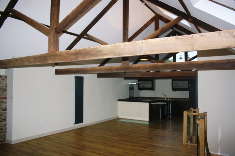 Spacious open-plan room with exposed wooden beams on the ceiling, light-colored walls, and wooden flooring. A modern kitchen with a black and white color scheme is visible in the background, along with a staircase and large windows.