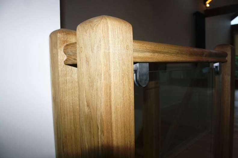 Close-up of a wooden staircase railing with a modern design. The railing features a smooth, light wood finish with a glass panel, providing a sleek and contemporary look. The background is softly lit, highlighting the railing's craftsmanship.