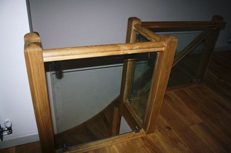 A staircase with glass panels and wooden frames. The wood is light-colored and polished, complementing the wooden flooring. The stairs lead downward, partially visible from the top.