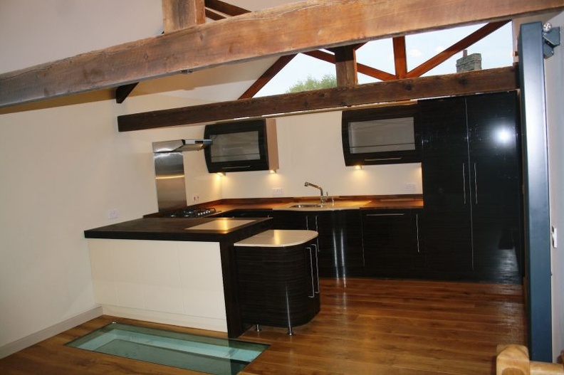 A modern kitchen with dark wooden cabinets, a glass panel in the floor, and exposed wooden beams. The kitchen features a corner counter with a cooktop, a sink, and overhead lights, set against a white ceiling and walls.