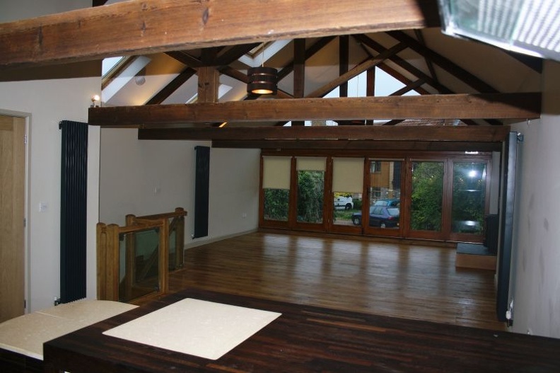 A spacious room with wooden beams and a high ceiling. The room has large windows with blinds that overlook trees and a parked car. The floor is wooden, and there's a wooden staircase railing on the left.