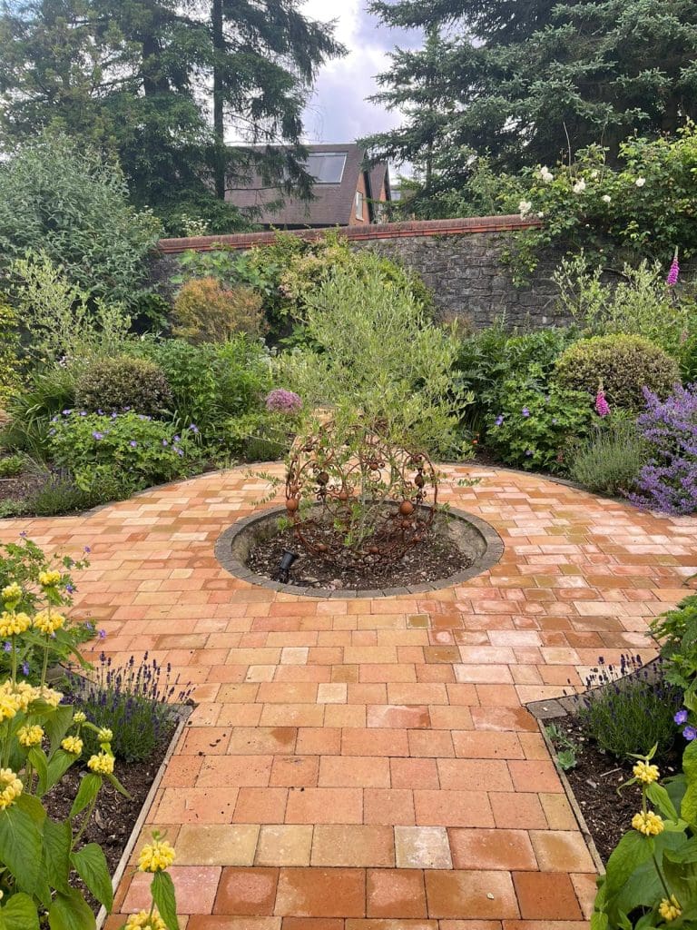 A garden with a circular brick pathway surrounding a small tree in the center. The path is bordered by lush greenery, purple flowers, and yellow blossoms. In the background, there's a stone wall and tall trees. Cloudy sky overhead.