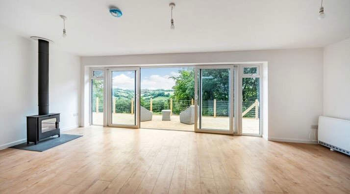 A spacious, bright room with wooden floors, a modern black fireplace, and large glass doors opening to a deck with outdoor seating. Beyond the deck is a scenic view of lush greenery and rolling hills under a clear sky.