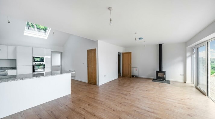 Spacious, modern open-plan living area with a light wooden floor. Features a sleek white kitchen with a skylight and granite countertops on the left, a central wood-burning stove, and glass sliding doors on the right leading to a balcony with a view.