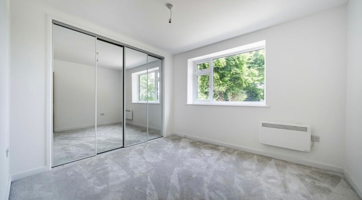 A bright, empty room with light grey carpet, white walls, and a large window on the right. There is a wall-mounted heater below the window and a closet with sliding mirrored doors on the left. A light fixture hangs from the ceiling.