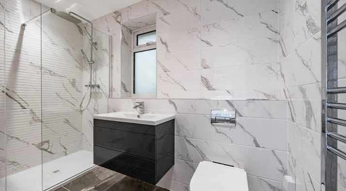 Modern bathroom with marble-patterned tiles, featuring a glass-enclosed shower on the left, a sleek sink with a black cabinet below the window, and a wall-mounted toilet beside a chrome towel rack on the right.