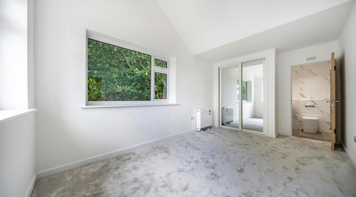 Minimalist bedroom with white walls and carpet. A large window reveals green foliage outside. A wooden door on the right leads to a bathroom with a tiled wall and visible toilet. Mirrored closet doors enhance the room's spacious feel.