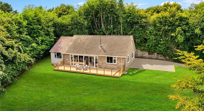 Aerial view of a small, single-story house with a sloped roof, surrounded by lush green trees and grass. The house has a deck with outdoor seating and large windows, set in a spacious yard under a clear blue sky.