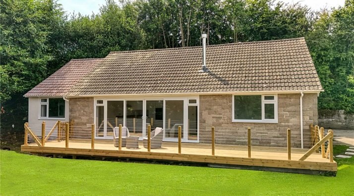 A single-story stone house with a sloped roof features large sliding glass doors and windows. A wooden deck with railing extends from the front, surrounded by a lush green lawn and trees in the background.