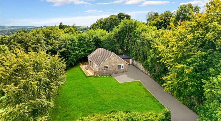 Aerial view of a small brick house surrounded by lush green trees and a large grassy lawn. A driveway leads to the house, which is nestled among dense foliage under a partly cloudy blue sky.