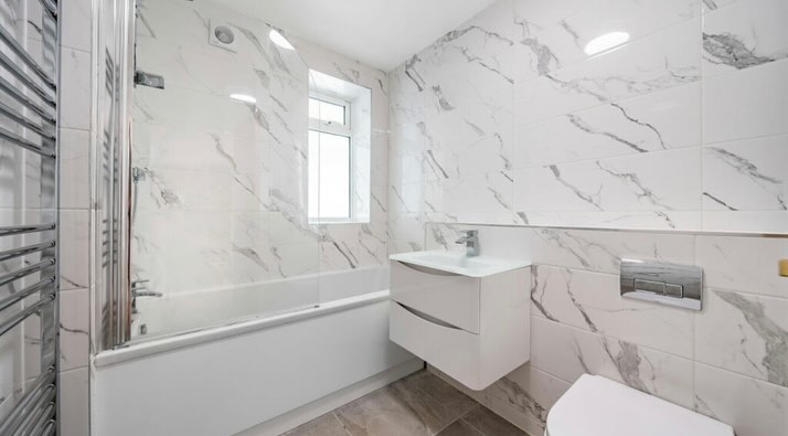 A modern bathroom with marble-patterned tiles, a white bathtub, a sleek white vanity with a sink, a wall-mounted toilet, and a large heated towel rail. A small window provides natural light.