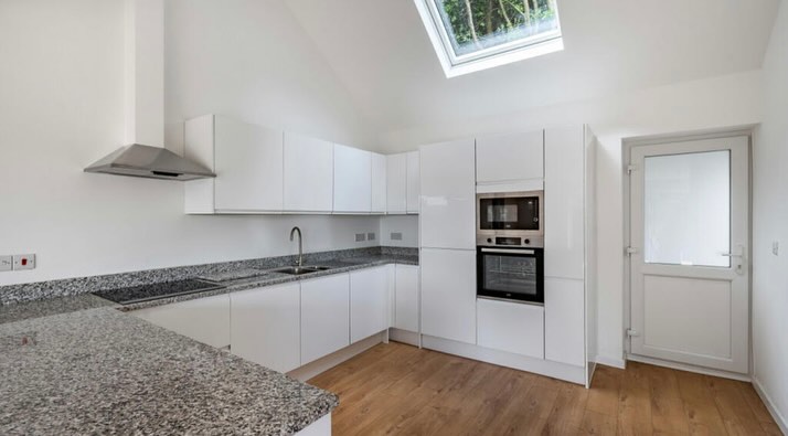 A modern kitchen with white cabinets, granite countertops, and a wood floor. There is an oven and microwave built into the cabinets, a stainless steel hood, and a skylight above. A glass door leads outside.