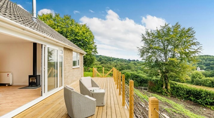 A wooden deck with two wicker chairs overlooks a lush, green landscape with trees and hills. The deck is attached to a white house with open sliding glass doors, revealing part of the interior with wooden flooring.