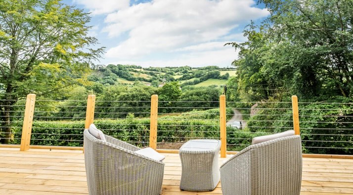 A serene outdoor deck with two wicker chairs and a small table overlooking a lush, green landscape. The view includes rolling hills and trees under a partly cloudy sky. The deck is bordered by a cable railing.