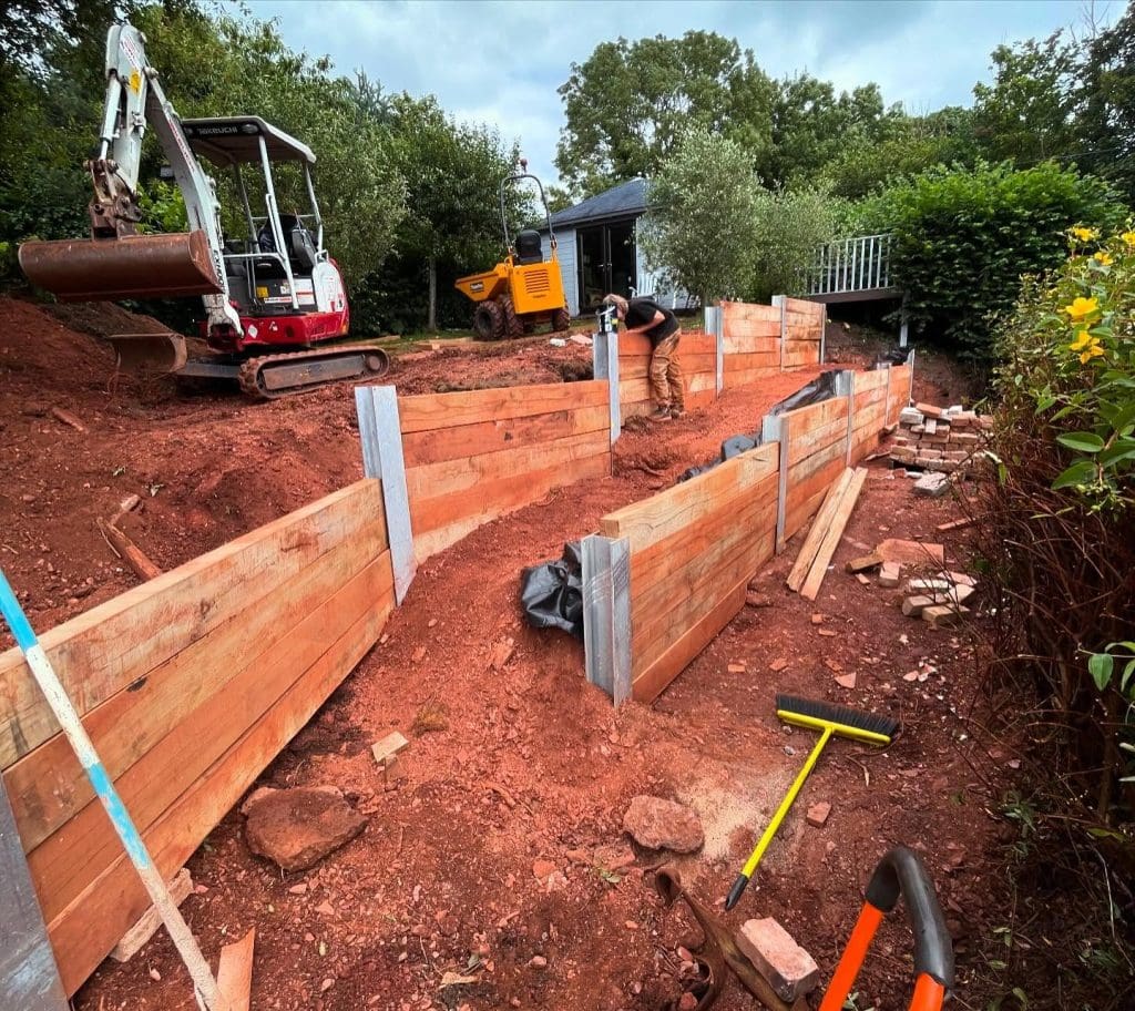 A construction site with a partially built wooden retaining wall on a sloped dirt path. Workers are seen operating machinery, including an excavator and a small bulldozer. Tools and construction materials are scattered around. Trees are in the background.