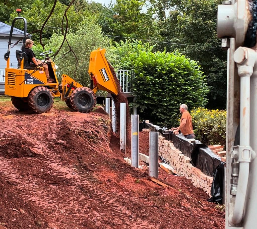 Two men work on a construction site. One operates a small yellow loader, dumping soil, while the other stands next to metal posts and a partially constructed brick wall. The setting is outdoors with trees and a house in the background.