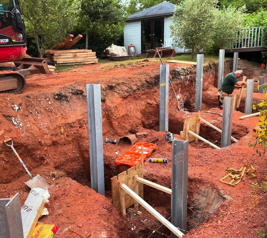 A construction site with steel beams inserted into deep red soil, supported by wooden braces. A person is working on the side, with tools and materials scattered nearby. A small building and trees are in the background.