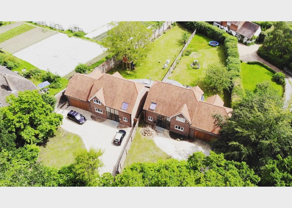 Aerial view of two adjacent brick houses with red roofs, surrounded by greenery. The houses have driveways with parked cars. A large lawn with a trampoline is visible in the background. Nearby, there are fields and other houses.