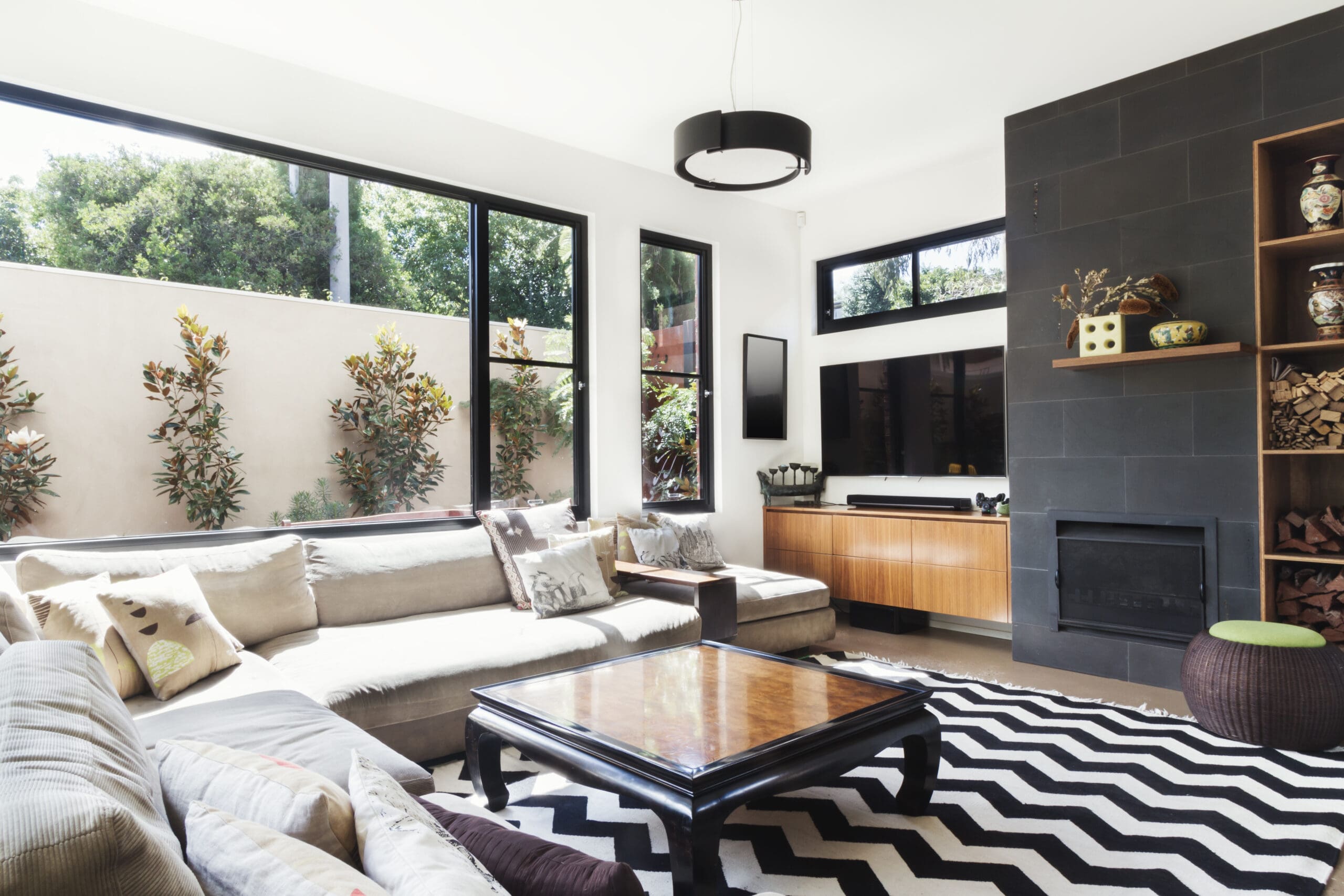 Modern living room with large windows, beige sectional sofa, and black-and-white zigzag rug. A wooden coffee table is centered, and a flat-screen TV is mounted above a wooden console. Shelves with decor are beside a black tiled fireplace.