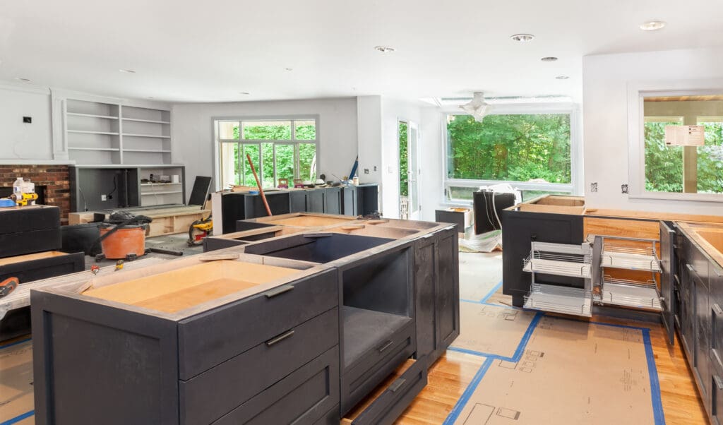 A kitchen under renovation with unfinished cabinets and shelves, raw wooden countertops, and construction tools scattered around. Large windows provide a view of green foliage outside, and protective flooring covers the area.