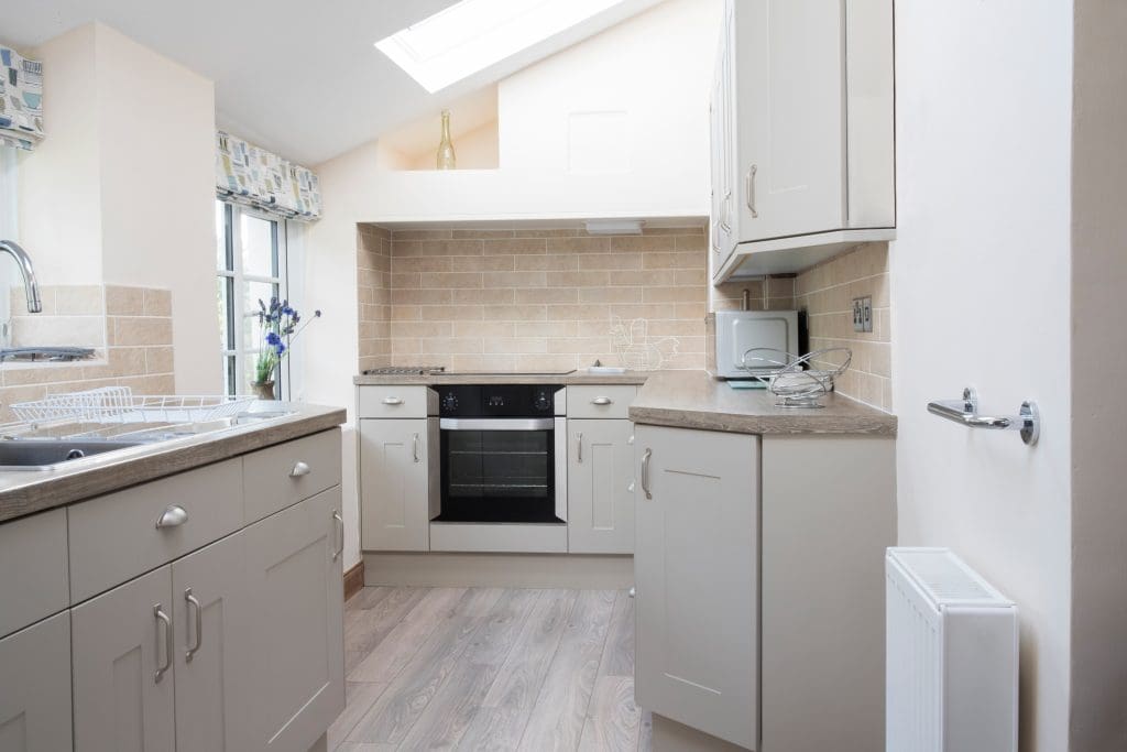 A small, modern kitchen with light gray cabinets, a black oven, and a beige tile backsplash. Natural light enters through a skylight. There is a microwave on the counter and a stainless steel sink with a drying rack, near a window.